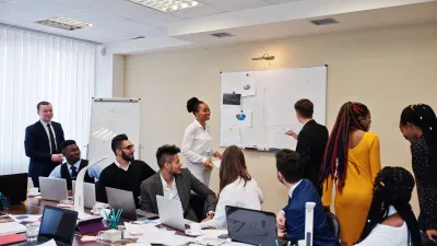 classroom with several professionally-dressed people working on laptops and two writing on a whiteboard