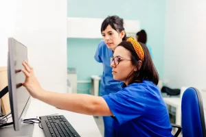 A female health worker analyses data on a computer screen whilst a colleague looks on.