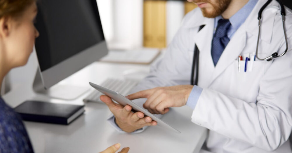 Unknown red-bearded doctor and patient woman discussing current health examination while sitting and using tablet computer in clinic, close-up. Medicine concept.